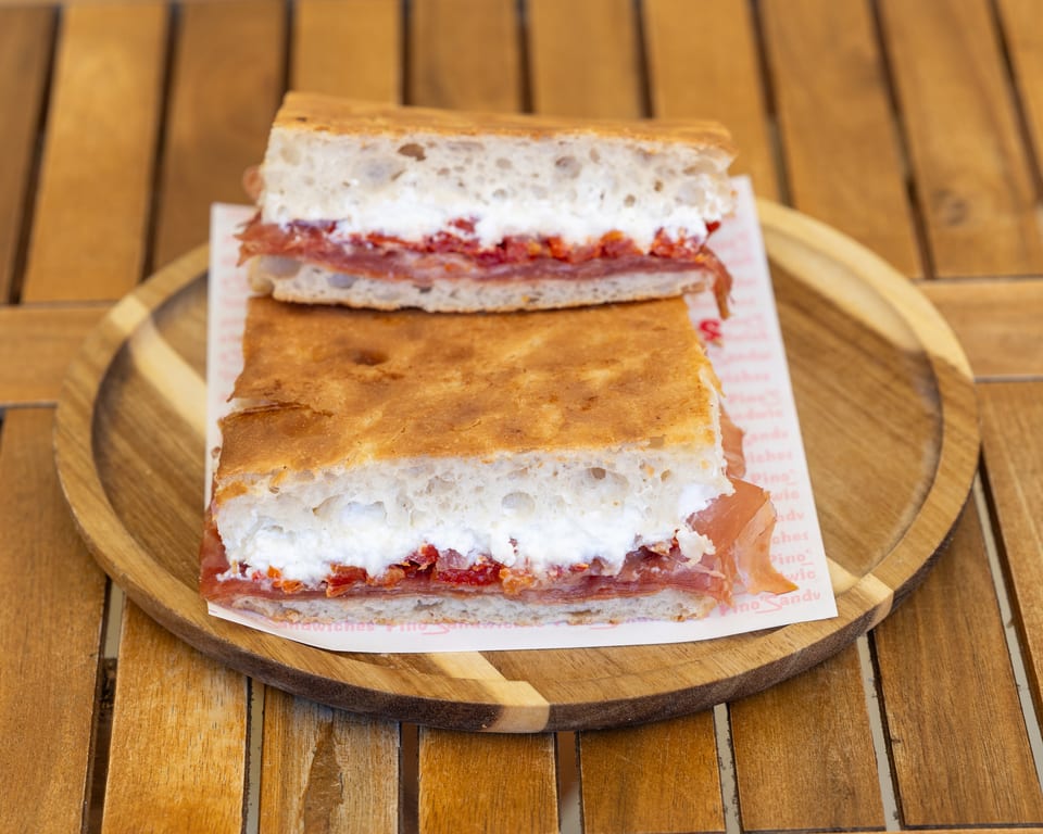 Two halves of a focaccia sandwich with prosciutto, ricotta, and roasted red peppers on a wooden plate.