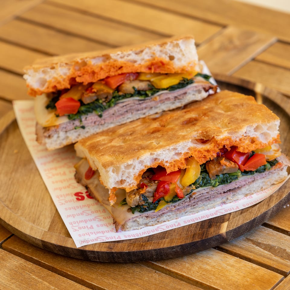 Two halves of a focaccia sandwich with meat, cheese, spinach, and roasted peppers on a wooden tray.