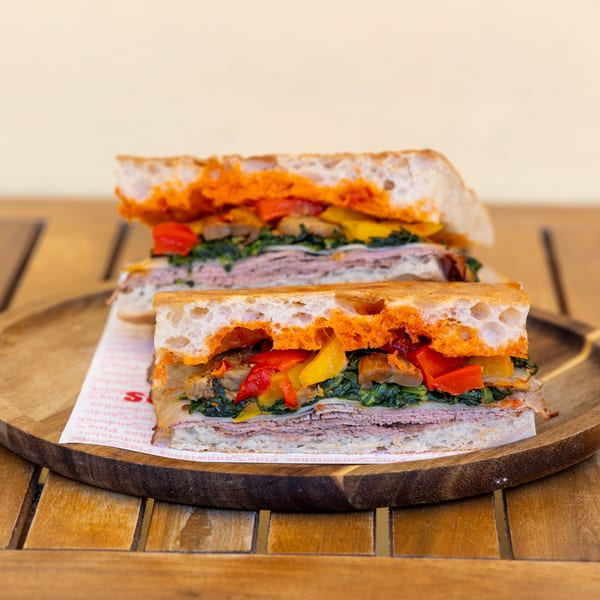 Two halves of a sandwich with meat, cheese, spinach, and roasted peppers on a wooden tray.