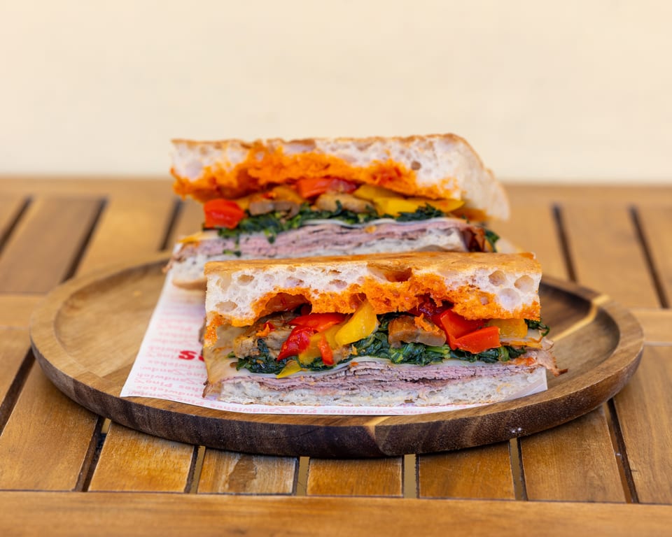 Two halves of a sandwich with meat, cheese, spinach, and roasted peppers on a wooden tray.