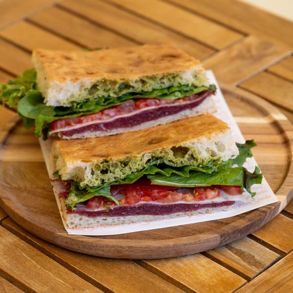 Two halves of a focaccia sandwich with pesto, lettuce, tomatoes, and meat on a wooden board.