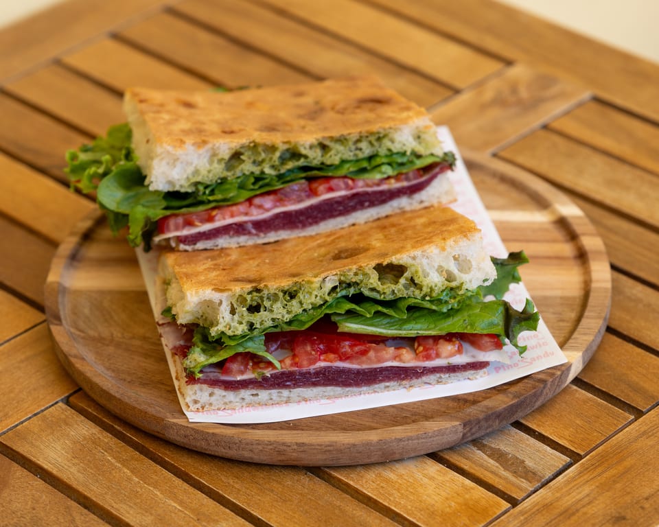 Two halves of a focaccia sandwich with pesto, lettuce, tomatoes, and meat on a wooden board.