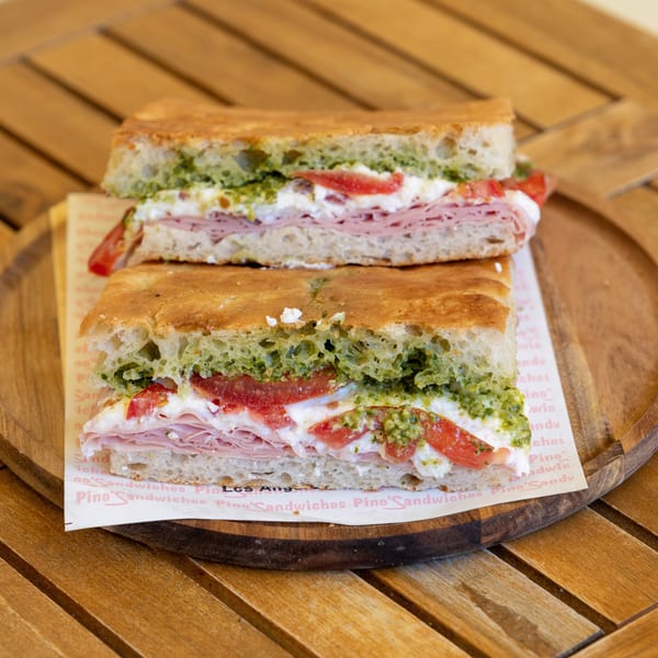 Two halves of a focaccia sandwich with ham, cheese, tomatoes, and pesto on a wooden tray.