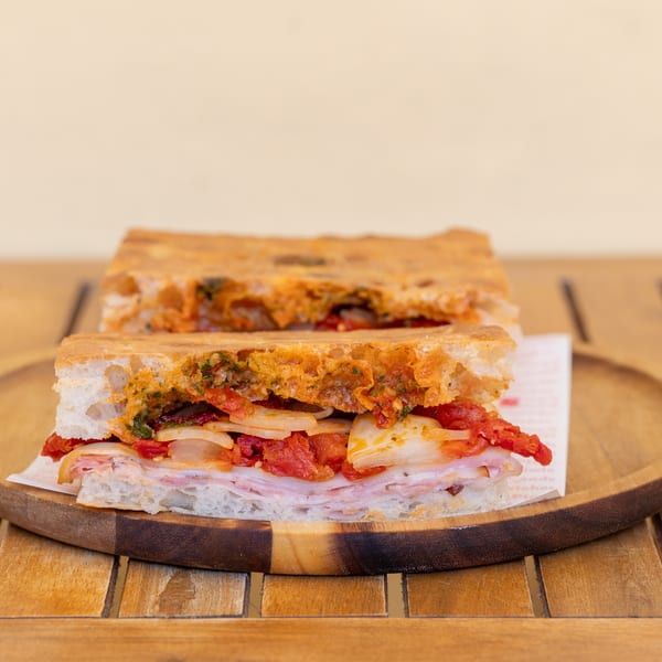 Two halves of a focaccia sandwich with ham, onions, and sun-dried tomatoes on a wooden plate.