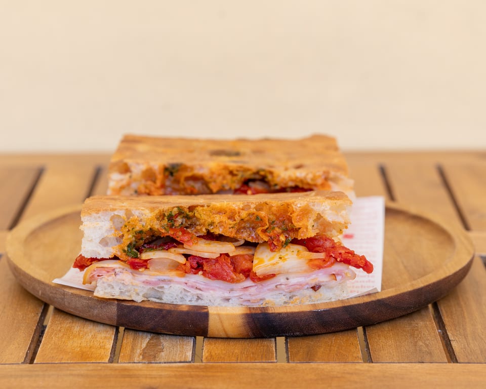 Two halves of a focaccia sandwich with ham, onions, and sun-dried tomatoes on a wooden plate.