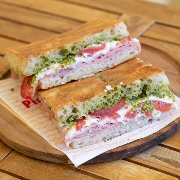 Two halves of a focaccia sandwich with ham, cheese, tomatoes, and pesto on a wooden tray.