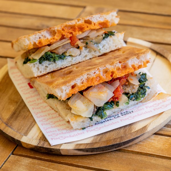 Two halves of a focaccia sandwich filled with greens, onions, and red sauce on a wooden plate.