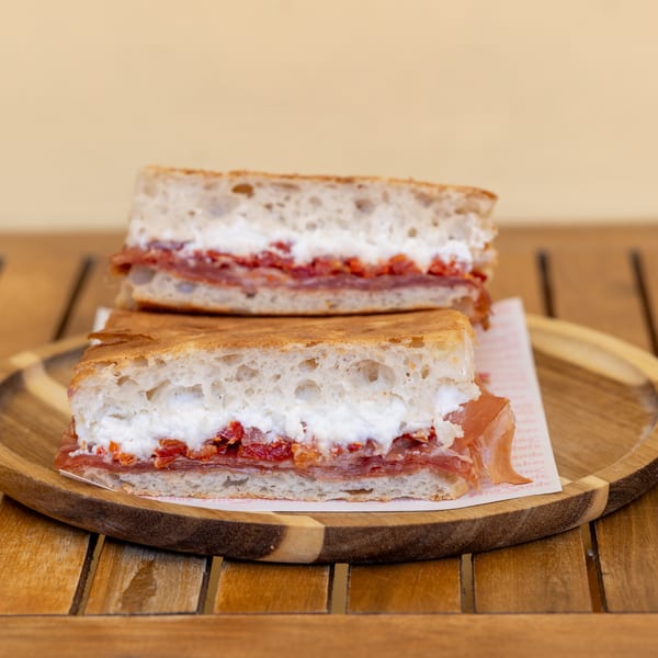 Two halves of a focaccia sandwich with prosciutto, ricotta, and sun-dried tomatoes on a wooden plate.