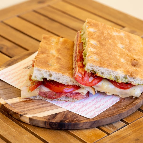 A Pino's Sandwich, cut in half, filled with salami, cheese, tomatoes, and pesto on focaccia.