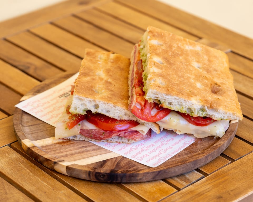 A Pino's Sandwich, cut in half, filled with salami, cheese, tomatoes, and pesto on focaccia.